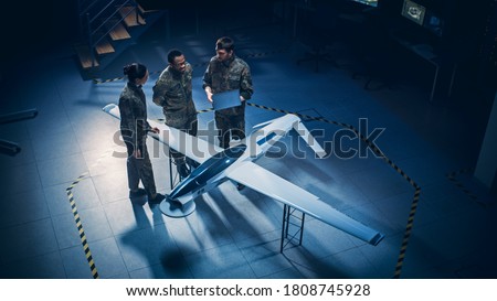 Army Aerospace Engineers Work On Unmanned Aerial Vehicle / Drone. Uniformed Aviation Experts Talk, Using Laptop. Industrial Facility with Aircraft for Performing Surveillance, Warfare Tactics, Attack