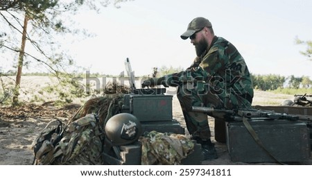The army commander sits behind a computer in the field, creating reports for the base, as a soldier approaches with a map. They discuss details while soldiers guard the camp in the background.