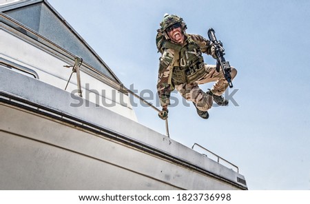 Army commando, special forces soldier equipped with body armour and battle helmet with radio headset, screaming and yelling while jumping from speed boat deck, landing on shore with assault rifle