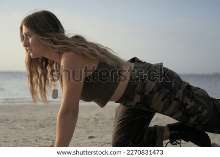 Army girl in olive drab crop top and camouflage pants crouching down on both hand and one knee. She appears to be doing a high crawl or resting between sets of pushups.