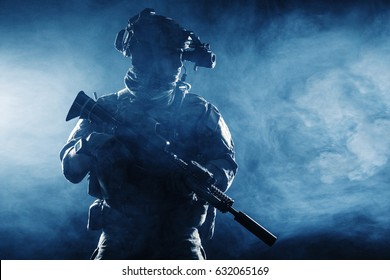 Army soldier in Combat Uniforms with assault rifle, plate carrier and combat helmet are on, Shemagh Kufiya scarf on his neck. Studio contour silhouette shot, backlight, dark glowing smoke background