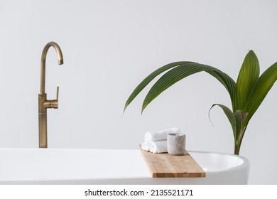 Aroma candle and folded towels at wooden shelf over contemporary tub. Modern copper faucet or water tap against copy space background. House decor at bright home with white interior design in bathroom