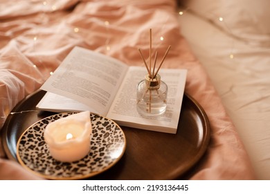 Aroma Sticks With Liquid Scented Perfume Water In Bottle With Burning Candle Staying On Open Paper Book On Wooden Tray In Bed Close Up. Aromatherapy. Home Cozy Atmosphere. 