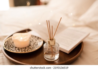 Aroma Sticks With Liquid Scented Perfume Water In Bottle With Burning Candle Staying On Open Paper Book On Wooden Tray In Bed Close Up. Aromatherapy. Home Cozy Atmosphere. 