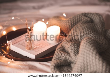 Aroma sticks with liquid scented water in bottle with burning candles staying on open book and knitted textile in bed closeup over lights at background . Winter holiday season. 