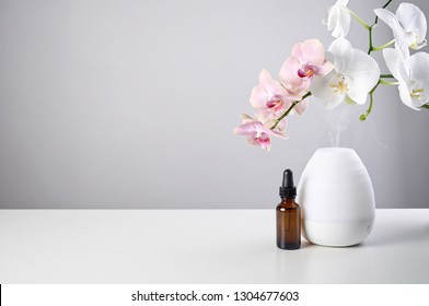 Aromatherapy at home. Ultrasonic Oil diffuser with glass amber bottle and orchid flowers on white table of gray background. Close up view with copy space