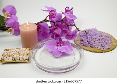 Aromatic bath salts, a bowl of water and a burning candle against the background of a branch of a blooming purple-pink orchid are on a white table