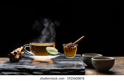 Aromatic herbal hot tea in glass teacup with steam and honey on black stone plate in dark background