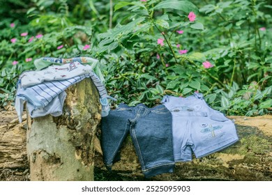 Arranged pile of baby clothes on tree trunk. The clothes are a blue and white striped onesie, a pair of blue denim jeans, and a blue and white striped shirt with an embroidered truck on the chest. - Powered by Shutterstock