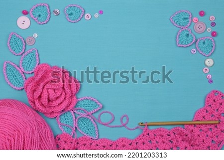 An arrangement of a pink rose, a hook, buttons, and a skein on a blue textile background.
