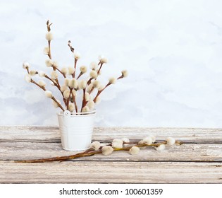 An arrangement of pussy willows in a small bucket on a rustic table top.