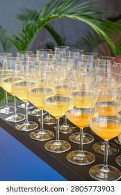 Array of elegant wine glasses filled with refreshing yellow and orange cocktails, arranged on a dark service table at an upscale outdoor event with tropical greenery in the background.