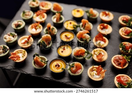 An array of small pastry shells filled with shrimp, purees, and herbs are neatly arranged on a black stone platter.