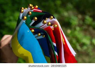 An array of tiny international flags arcs across a hand with the Ukraine banner nearest the viewer. Golden tips glitter while layered textiles show vivid color and movement against green bokeh