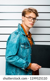 Arrested Hipster Man In Eyeglasses Holding Empty Prison Board And Looking At Camera In Front Of Police Line Up