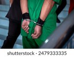 Arrested male criminal in prison uniform and handcuffs is brought by a Police officer to the Law and Justice Court for trial.