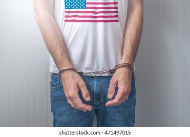 Arrested Man With Cuffed Hands Wearing Shirt With USA Flag. Unrecognizable Male Person In Jeans With Handcuffs Held In Police Station For Being Suspected Of A Crime.