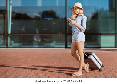 Arrival at Airport Concept. Beautiful smiling woman in hat using smart phone standing with baggage outdoors, full length - Powered by Shutterstock