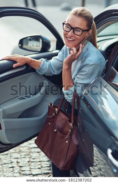 Arriving at work in the morning. Fashionable\
middle aged business woman with handbag getting out of her car, she\
is talking on phone and\
smiling