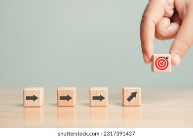 Arrow leading point to target icon on wooden blocks with businessman hand placing red goal dartboard symbol. Business agenda and mission setting concept. Dart board cube future vision strategy success