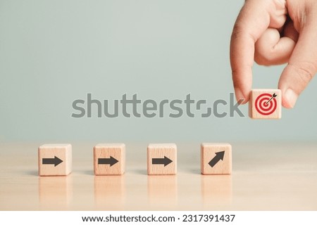 Arrow leading point to target icon on wooden blocks with businessman hand placing red goal dartboard symbol. Business agenda and mission setting concept. Dart board cube future vision strategy success