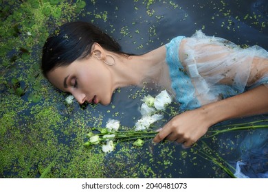 Art beautiful romantic woman lies in swamp in blue long dress with flowers. Portrait brunette in transparent dress in water swamp mud duckweed. Book cover - Powered by Shutterstock