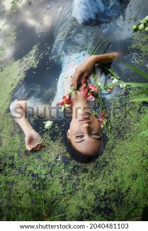Art beautiful romantic woman lies in swamp in blue long dress with flowers. Portrait brunette in transparent dress in water swamp mud duckweed. Book cover