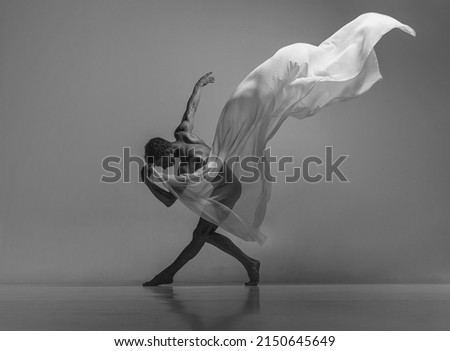 Art and beauty of male body. Black and white portrait of graceful muscled male ballet dancer dancing with fabric, cloth isolated on grey studio background. Grace, health, strength