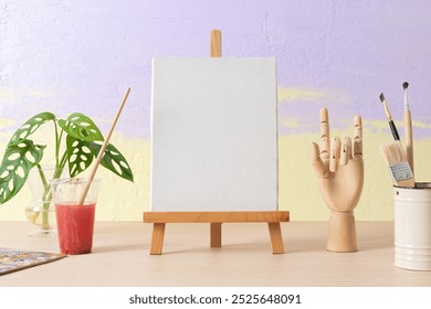 Art class with blank canvas on easel, surrounded by paintbrushes, paint, and a wooden hand model. Creative art tools and canvas for painting. Creative art workspace with blank canvas mockup - Powered by Shutterstock