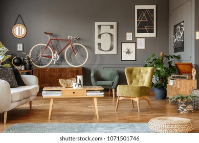 Art Collection On A Grey Wall, Wooden Coffee Table, Armchair And Red Bike In A Messy Living Room Interior