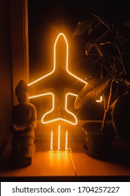 Kunstzusammensetzung von Buddha, Fikus und leuchtend mit gelber Neonlampe in Form von Flugzeug auf Sperrholzfensterscheibe.