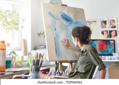 Art, creativity, hobby, job and creative occupation concept. Rear view of busy female artist sitting on chair in front of easel, painting with fingers, using white and blue oil or acrylic paint - Shutterstock ID 703964362