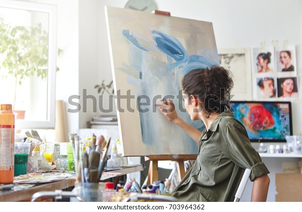 Art, creativity, hobby, job and creative occupation\
concept. Rear view of busy female artist sitting on chair in front\
of easel, painting with fingers, using white and blue oil or\
acrylic paint