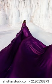Art fantasy beautiful woman posing in a purple purple flying dress, with a long train of fabric. Behind her is a winter frozen shore covered in blue and white ice. The model stands with her back