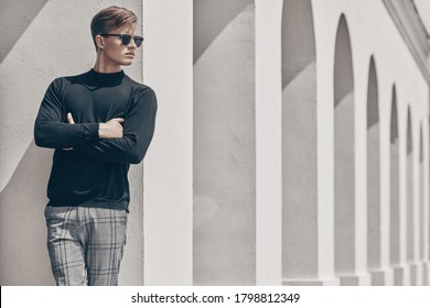 Art Fashion Portrait. Handsome Young Man Model In Black Pullover And Black Sunglasses Posing Next To White Columns In The Old Town. Men's Style, Beauty.