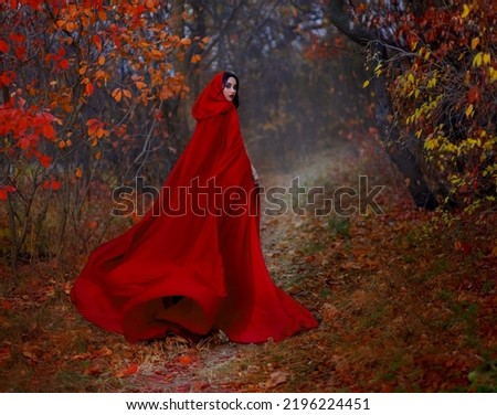 Art gothic fantasy woman like red riding hood walks in dark autumn forest. long silk cloak flutters, waving fly in wind fabric in motion. Girl princess looks back. Fallen orange leaves dark trees