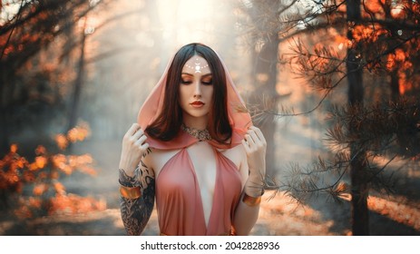 Art photo. Fantasy woman priestess pagan witch, moon sign on forehead. Lady elf in autumn forest. Dark trees background. Girl princess in orange silk dress, hood on head. Sexy red haired fashion model