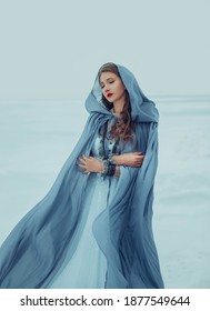 Art photo. Fantasy young woman fairy elf in blue cape with hood stands in cold wind. Winter nature background, white snow. Girl Queen walks in medieval dress, silk cloak, fabric is waving, fluttering.