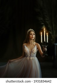 Art photo of medieval girl princess walks in dark gothic room. Woman queen is holding candlestick with burning candles in hand. Pink purple dress, long loose blonde hair, gold royal crown.