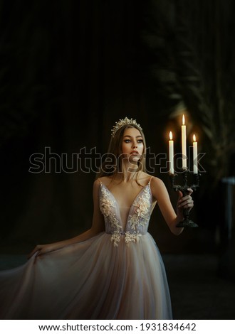 Art photo of medieval girl princess walks in dark gothic room. Woman queen is holding candlestick with burning candles in hand. Pink purple dress, long loose blonde hair, gold royal crown.