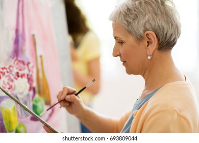 art school, creativity and people concept - happy senior woman artist with easel, paint brush and palette painting at studio - Powered by Shutterstock