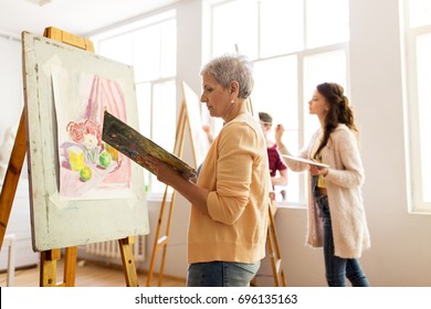 Art School, Creativity And People Concept - Happy Senior Woman Artist With Easel, Paint Brush And Palette Painting At Studio