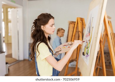 art school, creativity and people concept - student girl or young woman artist with easel, palette and paint brush painting still life picture at studio