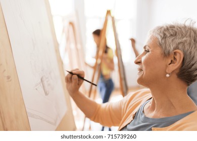 art school, creativity and people concept - happy senior woman artist with easel and pencil drawing picture at studio - Powered by Shutterstock