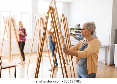 art school, creativity and people concept - happy senior woman artist with easel and pencil drawing picture at studio - Powered by Shutterstock