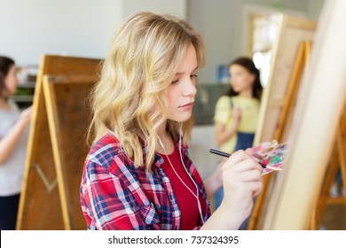 art school, creativity and people concept - student girl or artist with earphones, easel and paint brush painting at studio