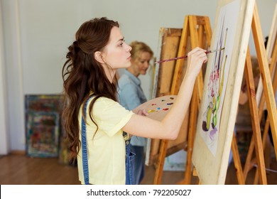 art school, creativity and people concept - woman with easel, palette and brush painting at studio