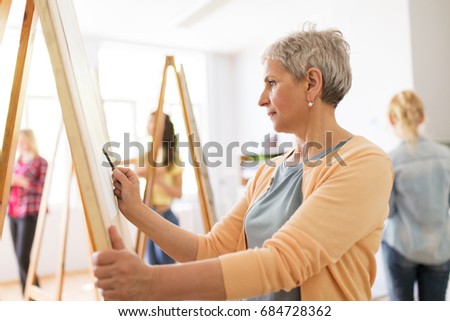 art school, creativity and people concept - happy senior woman artist with easel and pencil drawing picture at studio