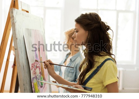 art school, creativity and people concept - woman with easel, palette and brush painting at studio