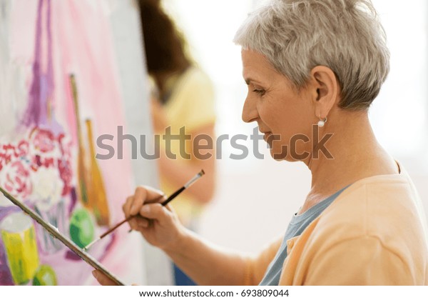 art school, creativity and people concept - happy\
senior woman artist with easel, paint brush and palette painting at\
studio